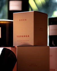 A box labeled ROEN Le Grand Topanga sits among other candle containers, with soft lighting and blurred green foliage in the background, suggesting the fresh earthy scent of this vegan soy wax candle.