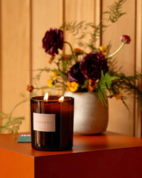 A lit Le Grand Topanga candle by ROEN in a dark glass jar rests on an orange surface, filling the air with a fresh earthy scent. In the background, a vase of flowers and greenery adds to the ambiance.