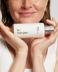 A smiling woman with wavy brown hair and a white tank top gently cradles a bottle of Rosie Jane Dulce Everyday Body Milk near her face, radiating the glow of nourishing moisturizer.