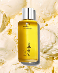 A yellow bottle of Dulce Everyday Body Oil by Rosie Jane, a hydrating vanilla-scented oil with a silver cap, sits on a creamy, textured background that looks like whipped body butter.