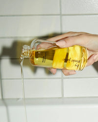 A hand with manicured nails pours Dulce Everyday Body Oil, a hydrating vanilla-scented oil by Rosie Jane, from a clear bottle against a white tiled background.