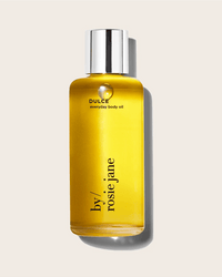 A clear bottle with a silver cap holds golden yellow Dulce Everyday Body Oil by Rosie Jane, hydrating and labeled on the front, set against a light beige background with a soft shadow.