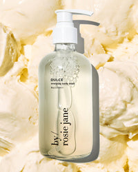 A clear pump bottle of Rosie Jane Dulce Everyday Body Wash sits on a creamy, textured off-white background that looks like swirled lotion or whipped cream.