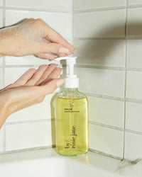 A hand pumps Rosie Jane’s Dulce Everyday Body Wash—creamy, hydrating cleanser with dulce fragrance—from a clear bottle by a white tiled tub, while another hand catches the soap suds.