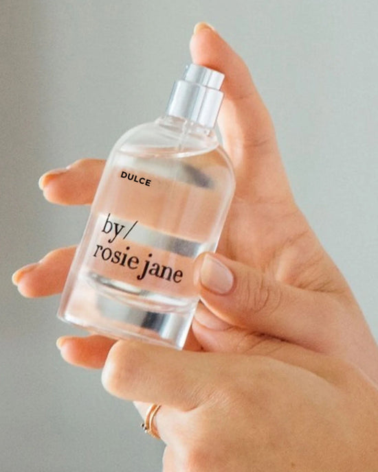 A hand holds a clear glass bottle of Rosie Jane Eau De Parfume in Dulce, a vanilla fragrance, against a neutral background.