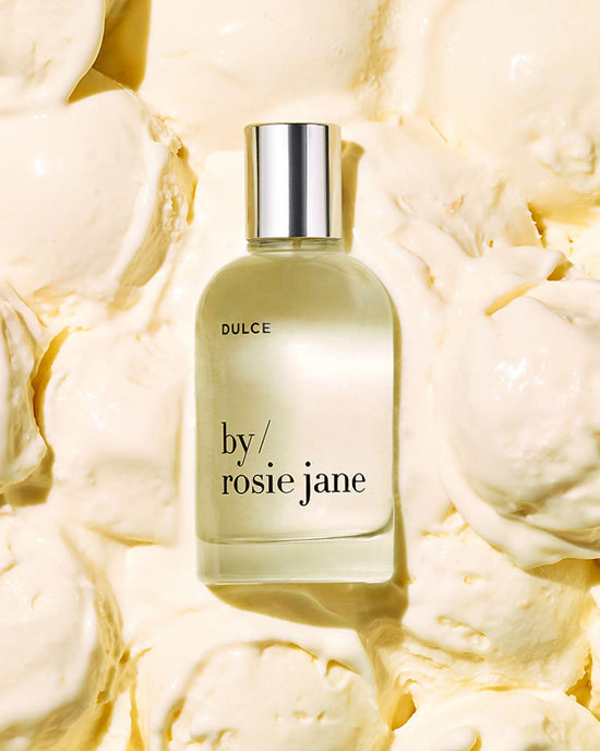 A clear glass bottle of Rosie Jane Eau De Parfume in Dulce with a silver cap sits on a creamy, textured yellow background.