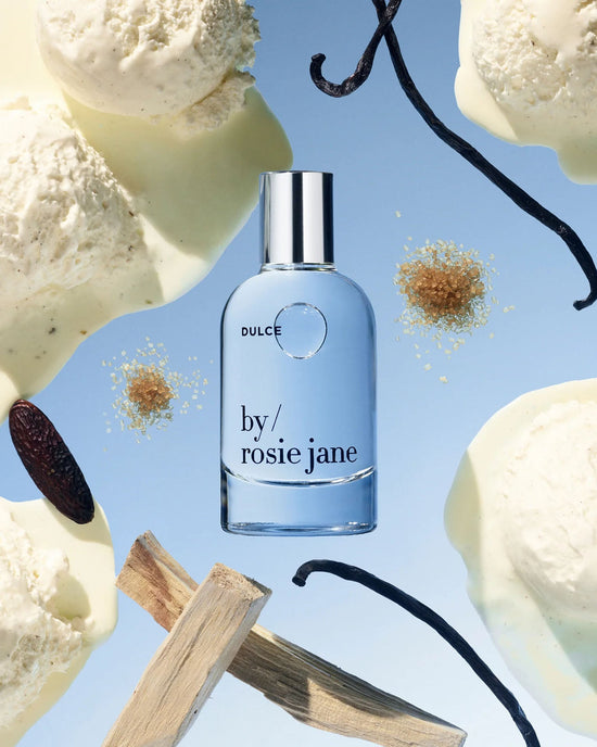 A bottle of Rosie Jane Eau De Parfume in Dulce sits among melting vanilla ice cream, vanilla beans, brown sugar, and wooden sticks on a blue background.