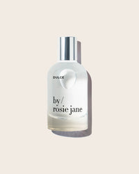 A clear glass bottle of Rosie Jane Eau De Parfume in Dulce with a silver cap, set against a light beige background.