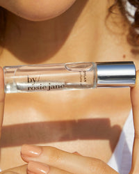 A person holds a clear glass bottle of Rosie Jane Perfume Oil in Dylan with a silver cap. Sunlight highlights their face, curly hair, and shoulders—capturing the perfect moment for this unisex fragrance.