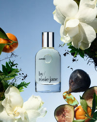 A bottle of Rosie Jane Eau De Parfume in James is surrounded by white flowers, fig, greenery, and an orange on a blue background.
