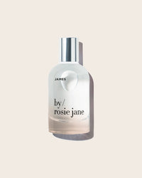 A clear glass bottle with a silver cap labeled Eau De Parfume in James by Rosie Jane stands against a light beige background, highlighting this signature fragrance from Rosie Jane.