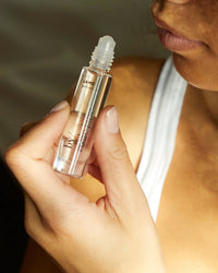 A person in a white tank top holds a clear glass Perfume Oil in James roller bottle by Rosie Jane near their lips, ready to apply the concentrated fragrance. The focus is on their hand, the bottle, and lower face.