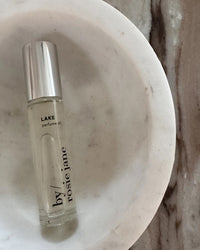 The Rosie Jane Perfume Oil in Lake features a concentrated fragrance in a glass bottle with a silver cap, displayed on a round white dish atop brown and gray-veined marble.