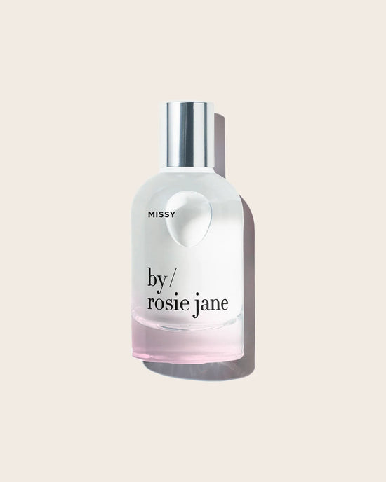 The Eau De Parfume in Missy by Rosie Jane features a clear glass bottle with a silver cap, light pink liquid, and notes of tropical fruits, displayed against a plain beige background.