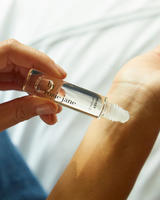 A hand applies Rosie Jane Perfume Oil in Missy from a glass roll-on bottle onto the wrist, with a soft white fabric background.