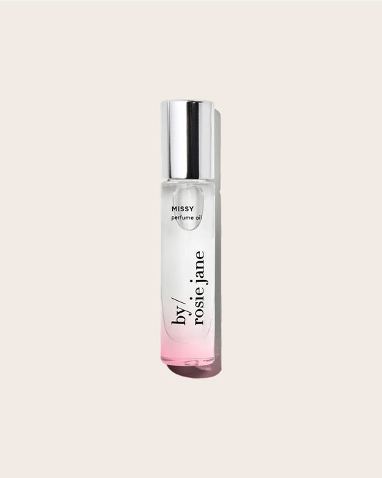 Rosie Janes Perfume Oil in Missy comes in a clear glass bottle with a silver cap, minimal black text, and a touch of pink at the base, reflecting its tropical scent, all set against a light beige background.