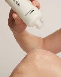 A person pours Missy Everyday Body Milk by Rosie Jane from a white bottle onto their knee, ready to apply this hydrating skincare product. The background is plain and neutral.