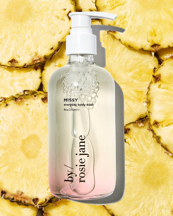 A clear pump bottle of Missy Everyday Body Wash by Rosie Jane rests on fresh pineapple slices, revealing a hint of light pink at the base. The cleanser offers gentle, hydrating care with a refreshing finish.