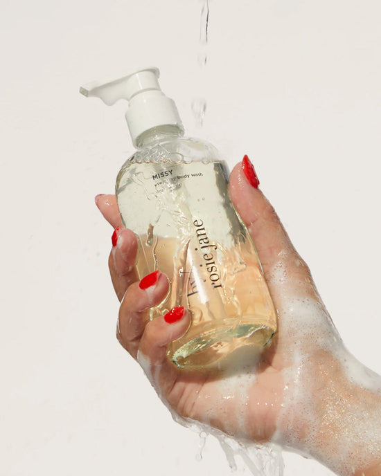 A hand with red-painted nails holds a clear pump bottle labeled Rosie Jane Missy Everyday Body Wash, a creamy hydrating cleanser covered in lather and water droplets against a plain light background.