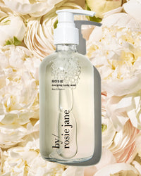 A clear pump bottle of Rosie Jane’s Rosie Everyday Body Wash sits on pale pink and white roses, displaying its transparent hydrating cleanser and elegant label, featuring a delicate white musk fragrance.