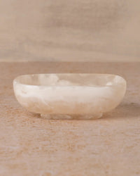The Saarde Flow Resin Soap Dish in Peach Blush features a shallow, oval shape with a smooth, polished surface and subtle marble finish, shown here on a beige textured background.