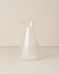The Saarde Kairos Angular Carafe in Clear, featuring vertical ribbed lines, rests on a neutral surface against a light background, accentuating its elegant design.