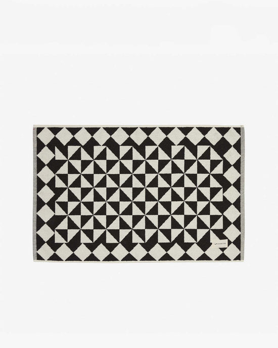 The Gatsby Bath Mat in Black by Slowtide features a black and white geometric pattern, is made from sustainable cotton, and has a small corner label.