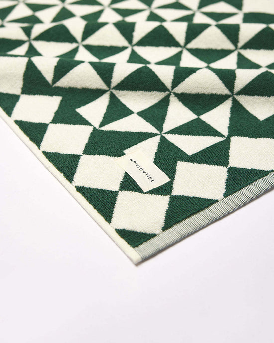 A close-up of the Gatsby Bath Mat in Sequoia by Slowtide, featuring a green and white geometric pattern on ultra-soft, sustainably sourced cotton, with a small Slowtide tag on the corner.
