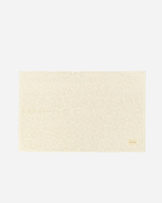 The Ginny Bath Mat by Slowtide is a rectangular, cream-colored mat made from absorbent, sustainably sourced cotton with a subtle floral pattern and a small label in the bottom right corner, shown against a plain background.