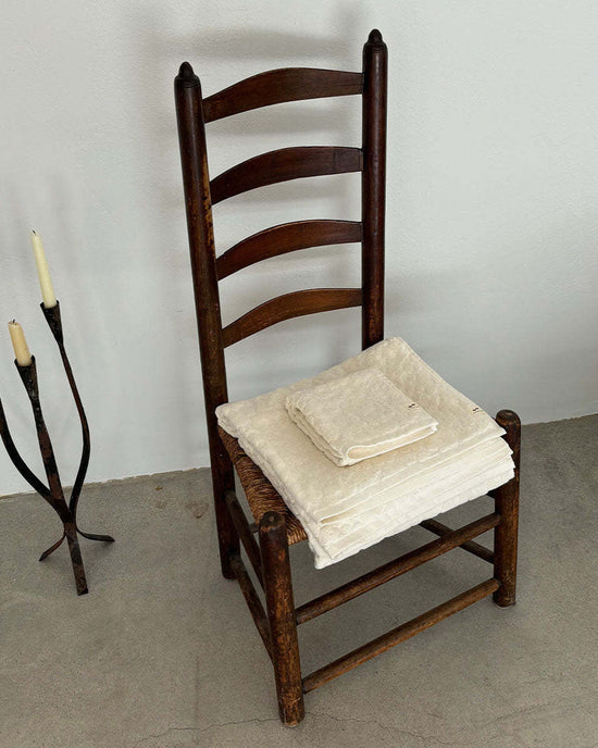 A wooden ladder-back chair with folded Slowtide Ginny Bath Towels on the seat sits on a concrete floor beside a metal candle holder with two candles.