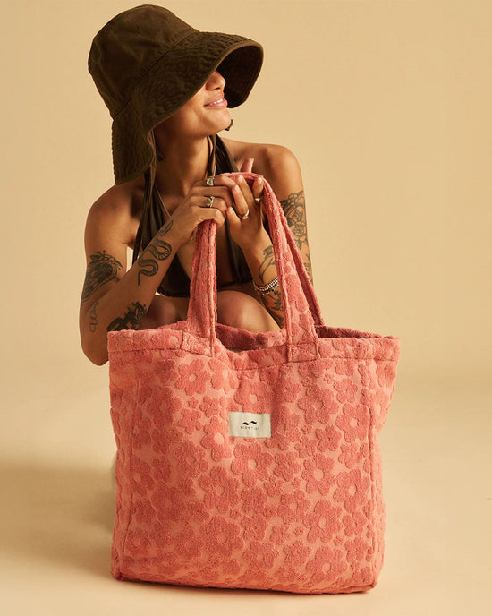 A person kneels against a beige background, holding the Slowtide Ginny Terry Tote Bag in Guava—crafted from sustainably sourced cotton and featuring a white label—while wearing a brown bucket hat and dark bikini top.