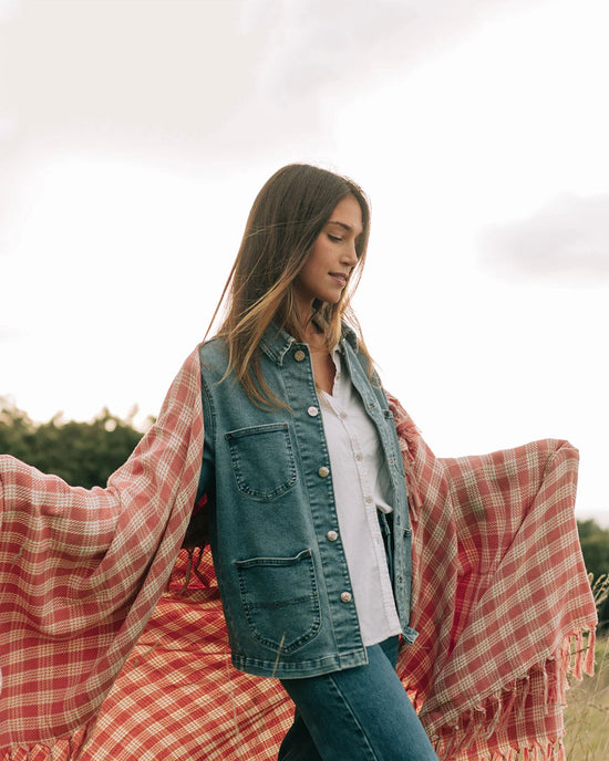 A woman stands outdoors with arms extended, draped in the Slowtide Palaka L/W Beach Blanket in Guava over a denim jacket, white shirt, and jeans.