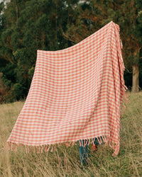 The Slowtide Palaka L/W Beach Blanket in Guava, featuring a large pink and white checkered design, is displayed outdoors in a grassy field with trees in the background, partially concealing the person holding it.