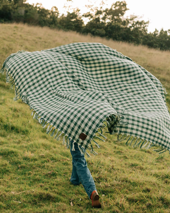 A person walks on grass holding the Slowtide Palaka L/W Beach Blanket in Sequoia, a large green and white checkered blanket that billows and covers most of their body.