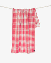 The Slowtide Palaka Woven Beach Towel in Guava, featuring a pink and white plaid pattern, hangs on a white rope against a plain white background.