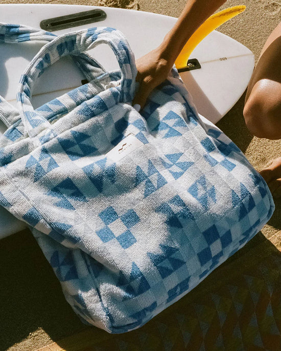 A person sets the Slowtide Dream Terry Tote Bag in Lagoon, crafted from sustainably sourced cotton, on a surfboard at the beach.