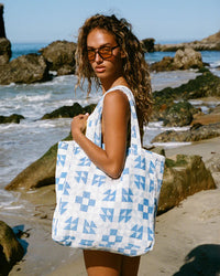 A woman with curly hair and sunglasses stands on a rocky beach holding Slowtides The Dream Terry Tote Bag in Lagoon, crafted from sustainably sourced cotton.