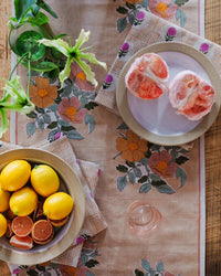 A Dia Table Runner in Cafe au lait by Soil to Studio decorates the table, which holds a plate of lemons, halved grapefruit, a glass, and a vase with greenery.