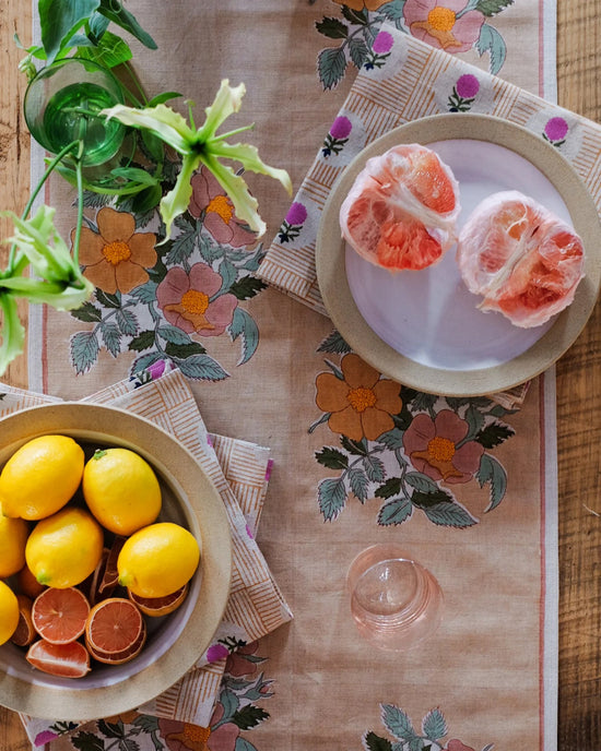 A Dia Table Runner in Cafe au lait by Soil to Studio decorates the table, which holds a plate of lemons, halved grapefruit, a glass, and a vase with greenery.