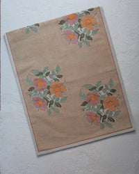 The Soil to Studio Dia Table Runner in Cafe au lait from the Valley of Flowers Collection showcases a beige linen base with a pink, orange, and yellow floral pattern and green leaves, bordered and laid flat on a textured white surface.