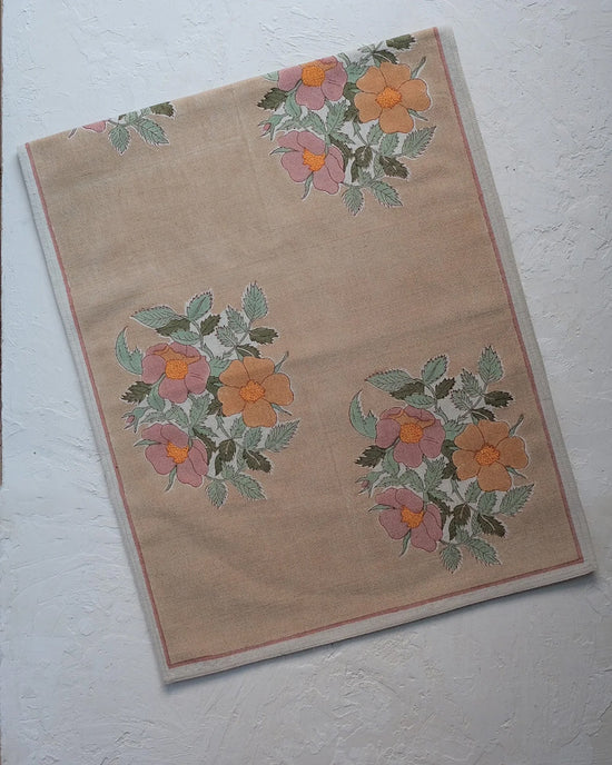 The Soil to Studio Dia Table Runner in Cafe au lait from the Valley of Flowers Collection showcases a beige linen base with a pink, orange, and yellow floral pattern and green leaves, bordered and laid flat on a textured white surface.