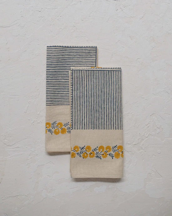 Two Mehak Table Napkins in Daffodil by Soil to Studio, featuring beige cotton with vertical navy stripes and a yellow floral pattern inspired by the Valley of Flowers, are set on a light textured surface.