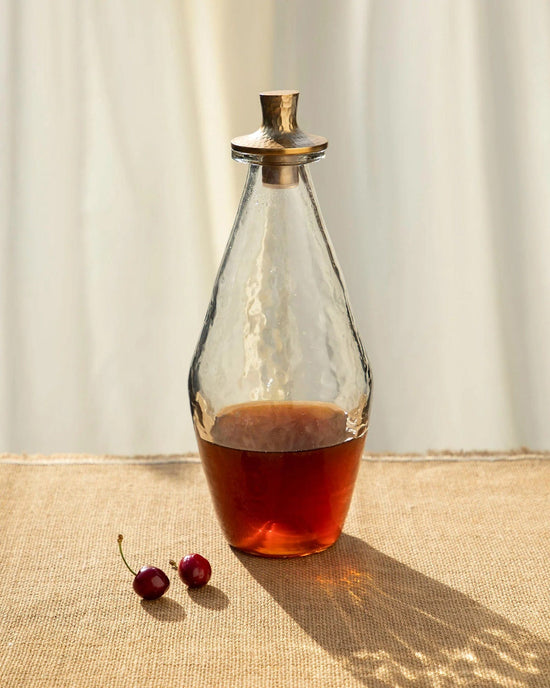 The Collectives Pebbled Decanter Classic, featuring a hammered aluminum stopper and amber liquid inside, sits on burlap beside two cherries with soft cream curtains in the background.