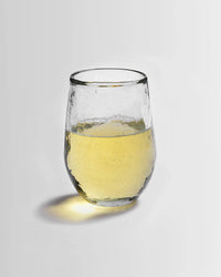 A Pebbled Small Glass from The Collective, filled halfway with pale yellow liquid, rests on a plain white surface and casts a slight shadow to the left.