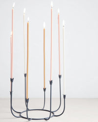The Floral Society’s Beacon Long Stem Candelabra in Smoke, featuring eight lit pastel taper candles, is showcased against a plain light background.