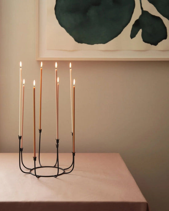 The Floral Society’s Beacon Long Stem Candelabra in Smoke, featuring seven lit taper candles, rests on a pink tablecloth beneath a framed abstract green artwork on the wall.