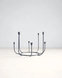 The Floral Societys Beacon Long Stem Candelabra in Smoke features six upward-curving arms with flower-shaped holders, crafted from minimalist black wrought iron and displayed on a light surface against a plain background.