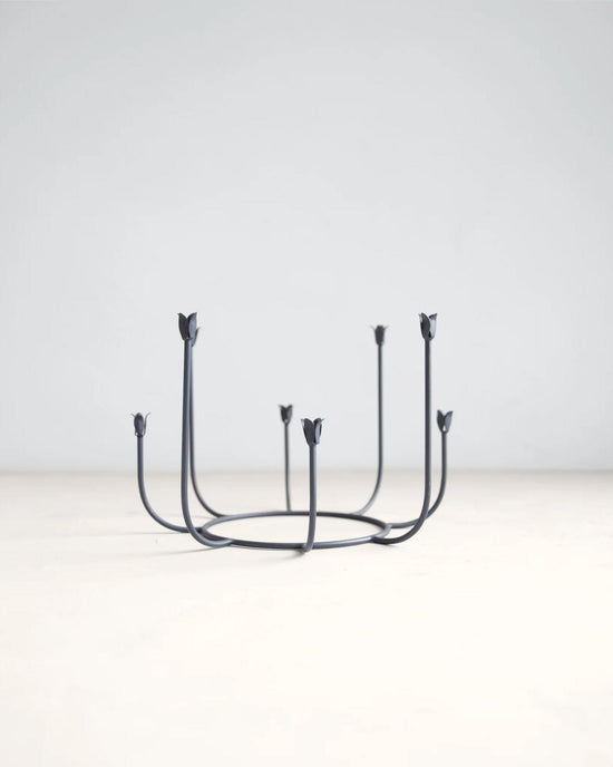 The Floral Societys Beacon Long Stem Candelabra in Smoke features six upward-curving arms with flower-shaped holders, crafted from minimalist black wrought iron and displayed on a light surface against a plain background.