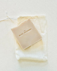 A beige bar of Coconut - Handmade Soap by The Quiet Botanist, embossed with the brand name, rests on a white fabric drawstring pouch atop a light surface.