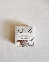 A square box labeled Coconut - Handmade Soap by The Quiet Botanist rests on a light textured surface, decorated with floral designs and showcasing its natural coconut shea butter soap inside.